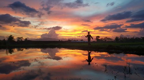 Silhouette person standing by lake against sky during sunset