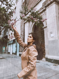 Young woman looking away while standing against tree in city