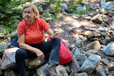 Senior woman sitting on the rock in nature and eating sandwich