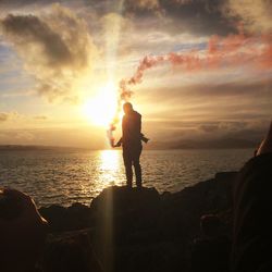 Man holding distress flare while standing on rock against sea during sunset