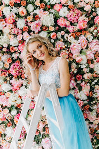 Young woman wearing blue dress standing on ladder against flowering plants