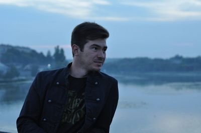 Young man looking away while standing against lake during sunset