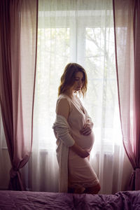 Pregnant girl in a pink silk nightie stands against the background of a large window with curtains
