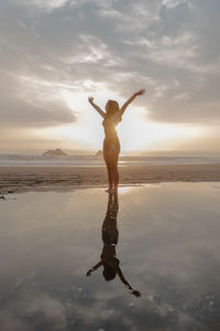 Full length of teenage girl with arms raised standing at beach against sky during sunset