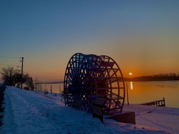 Ferris wheel by snow covered land against sky during sunset