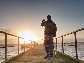 Rear view of man standing by railing against sky during sunset