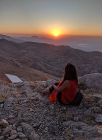 Rear view of woman sitting on rock against sky during sunset