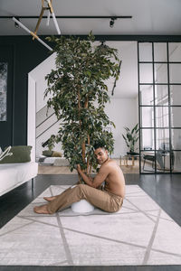 Shirtless man with eyes closed embracing houseplant in living room at home