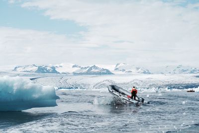 Rear view of man standing in boat on sea against cloudy sky during winter