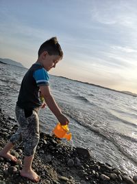 Boy playing with toy in sea against sky during sunset