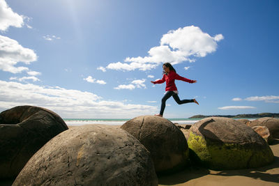 Woman jumping over rocks at beach against sky