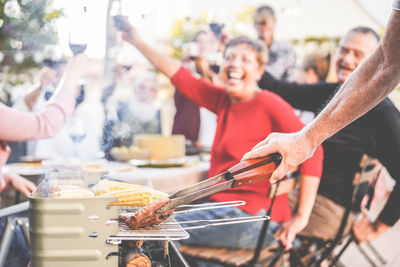 Cropped hand of man preparing meat on barbecue with cheerful friends in background