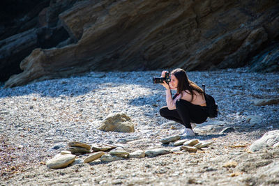 Beautiful woman crouching taking a picture of a pebbles beach at a creek side shot person