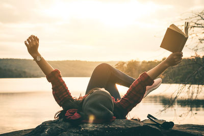 Woman reading book while lying on lakeshore during sunset