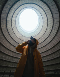 From below young male in hoodie touching face and looking up while standing under hole in ceiling inside abandoned industrial facility