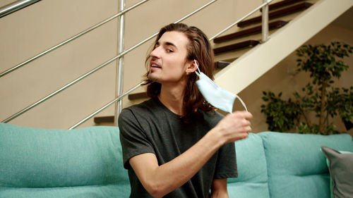 Young man removing mask while sitting on sofa at home