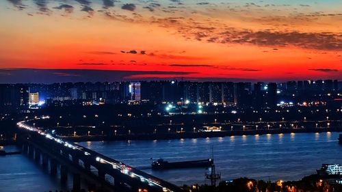 Illuminated city by sea against sky at sunset