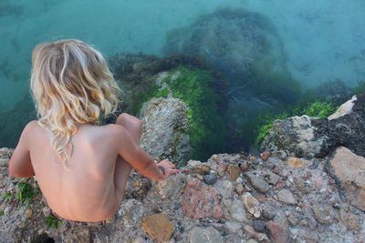 Rear view of shirtless boy crouching on cliff over sea