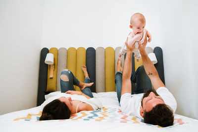 Father pretending that playful baby flying like superhero while chilling together with wife and kid on soft bed at home