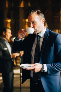 Mature businessman drinking coffee while standing against reflection of male colleague on glass