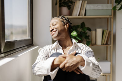 Smiling woman enjoying sunlight sitting by window at home