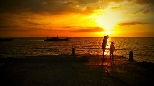 Woman and child on pier by sea against orange sky during sunset