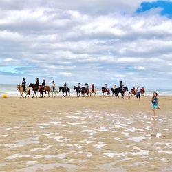 People riding horses at beach against cloudy sky