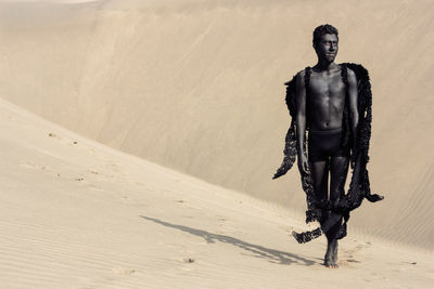 Full length of shirtless model with costume wing standing at desert