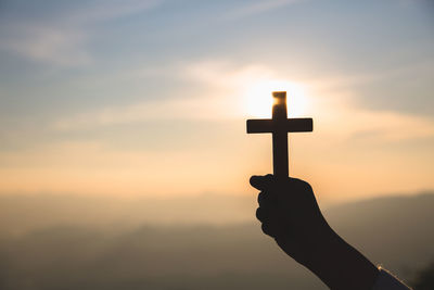 Silhouette hand holding cross against sky during sunset