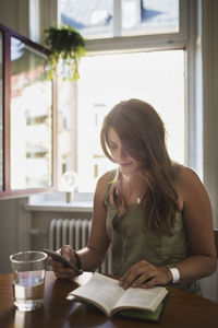 Young woman using smart phone while reading travel book at home