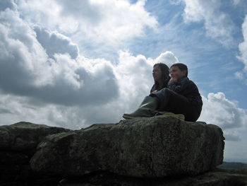 Low angle view of mother with son sitting on rock against cloudy sky