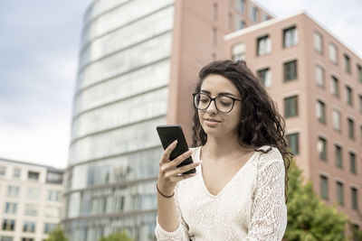 Smiling woman holding smart phone sitting against building