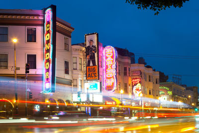 Light trails on road by buildings against sky at night