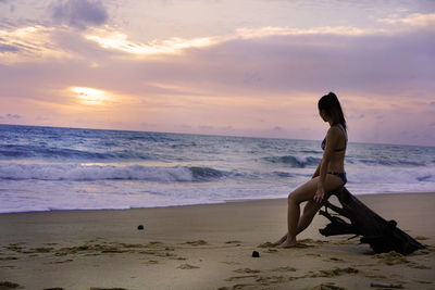 Girl on driftwood at beach against sky during sunset