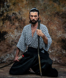 Portrait of samurai with sword kneeling against weathered wall