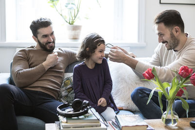 Father is making daughter wear eyeglasses while sitting with partner on sofa at home