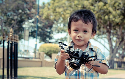 Portrait of cute boy holding camera while standing against trees