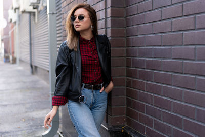 Young woman wearing sunglasses smoking cigarette while standing against brick wall