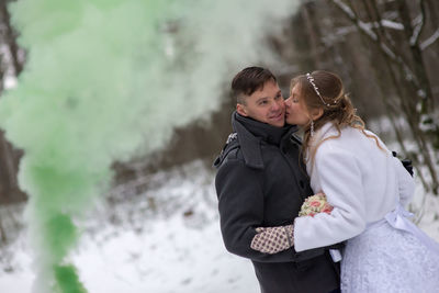 Bride kissing groom while standing at forest during winter