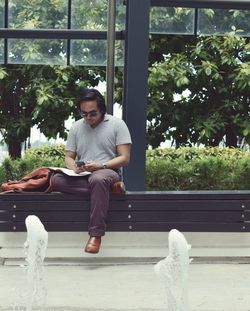 Young man sitting using phone while sitting against modern glass building