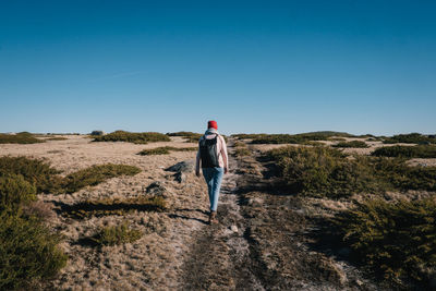 Rear view of young woman with backpack walking on landscape against clear blue sky