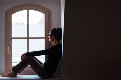 Side view of woman looking through window while sitting by wall