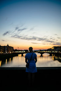 Rear view of woman standing over river against sky during sunset