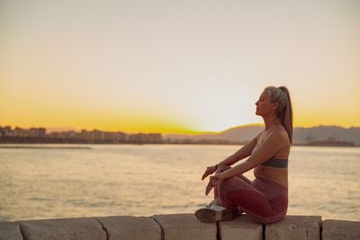 Side view of woman sitting on retaining wall against sky during sunset