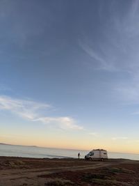 A lone person next to their camper van against a scenic view of sea against sky during sunset. 