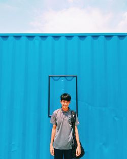 Portrait of teenage boy with backpack standing against blue wall during sunny day