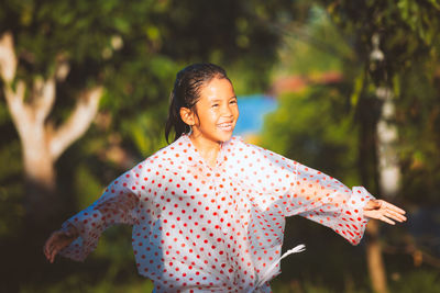 Happy girl wearing raincoat with arms outstretched standing in park during rainfall