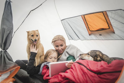 Mother and daughter watching digital tablet while lying with pets in tent at camping site