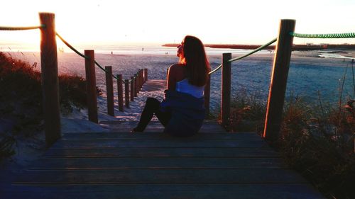Woman sitting on steps at beach against sky during sunset