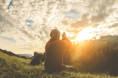 Rear view of mid adult man meditating while sitting on field against cloudy sky during sunset